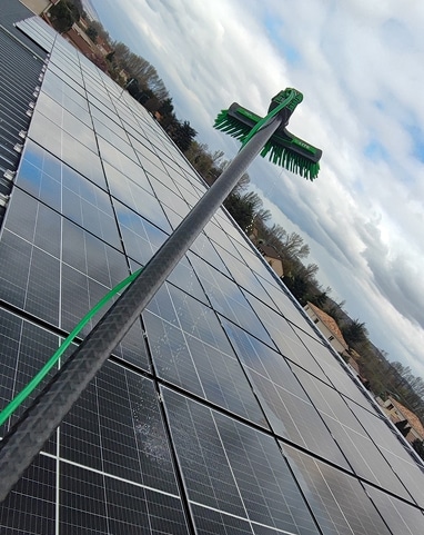 Nettoyage de panneaux solaires avec une longue perche télescopique et une brosse verte, sous un ciel nuageux, vue en plongée sur le toit.