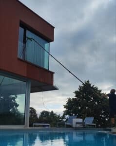 Nettoyage vitres haute perche près piscine moderne Homme utilisant une perche télescopique pour nettoyer la fenêtre d'un bâtiment moderne avec piscine en contrebas, par temps nuageux.