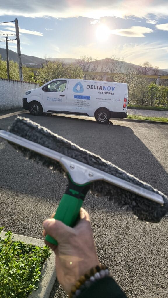 Un laveur de vitres tient un manche avec une tête de lavage devant un fourgon blanc DELTANOV NETTOYAGE (Tél : 06 62 93 46 62) au soleil.