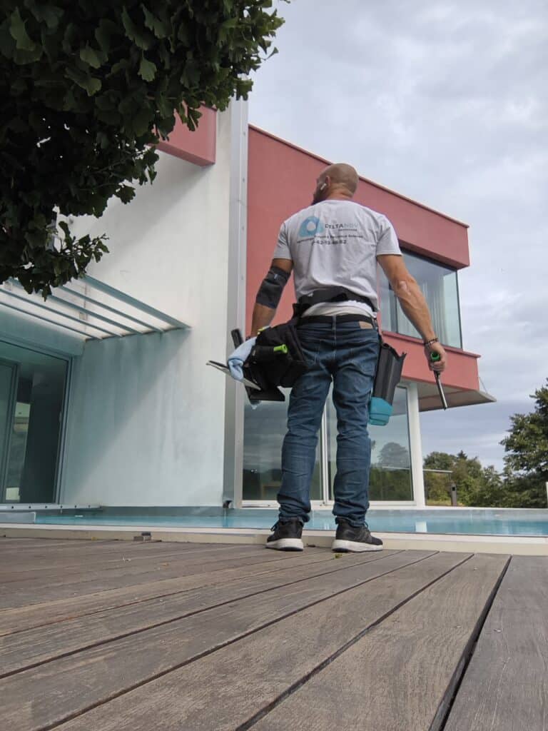 Professionnel DELTANOV "Nettoyage Vitres & Panneaux Solaires" sur une terrasse en bois. Il tient ses outils face à la façade vitrée de la maison moderne.