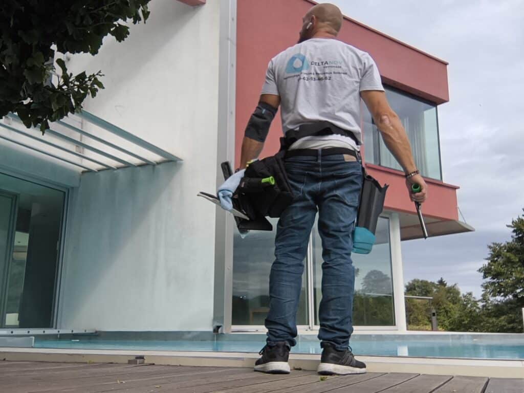 Professionnel DELTANOV "Nettoyage Vitres & Panneaux Solaires" sur une terrasse en bois. Il tient ses outils face à la façade vitrée de la maison moderne.