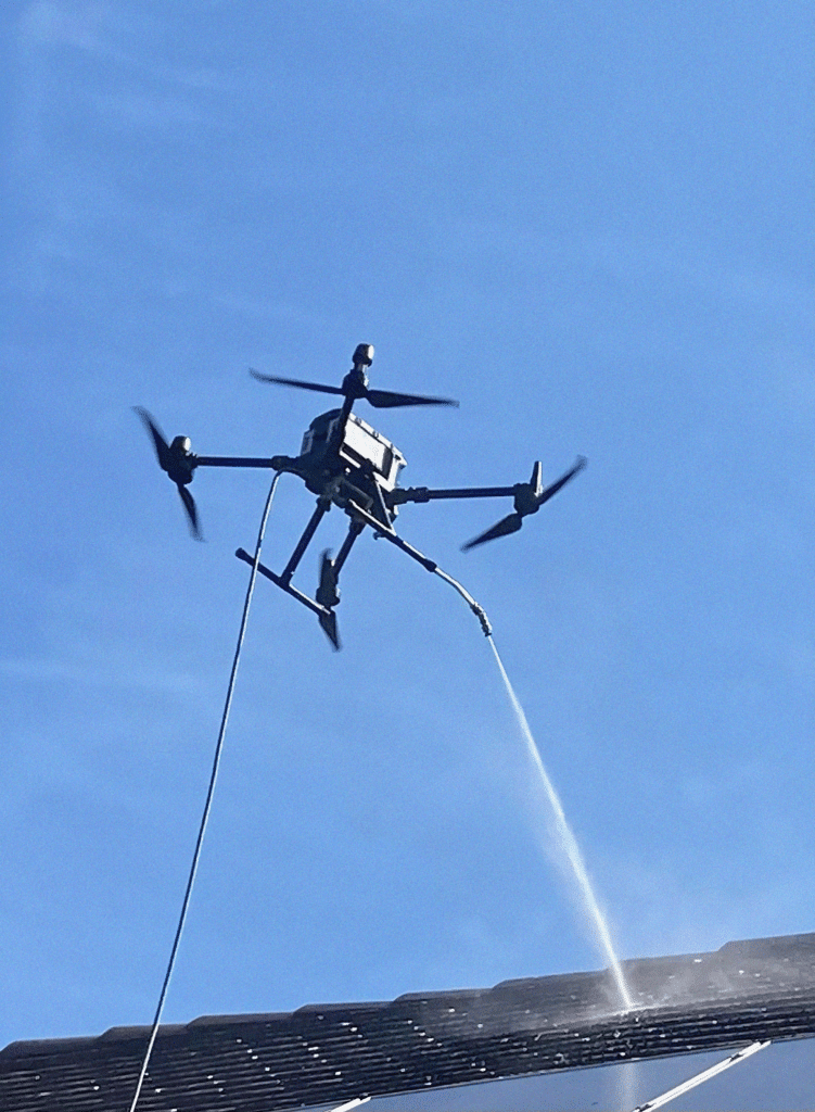 Drone de nettoyage pulvérisant de l'eau sur une toiture pour entretien, sous un ciel bleu dégagé.